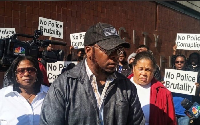 Amir Shakur speaking at the press conference with Samaria Rice beside him; 'No Police Brutality' signs and NBC News cameras behind them.